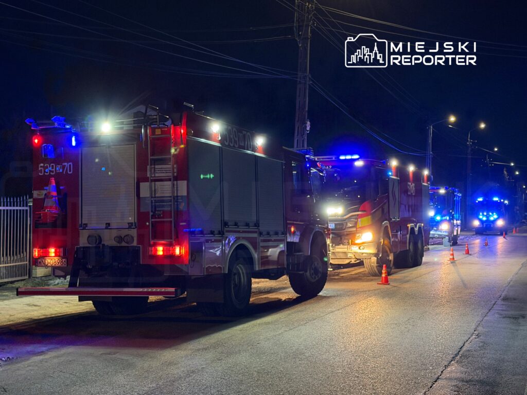 Karetka strażacka z włączonymi sygnałami świetlnymi stoi na ulicy nocą, otoczona przez inne pojazdy ratunkowe.