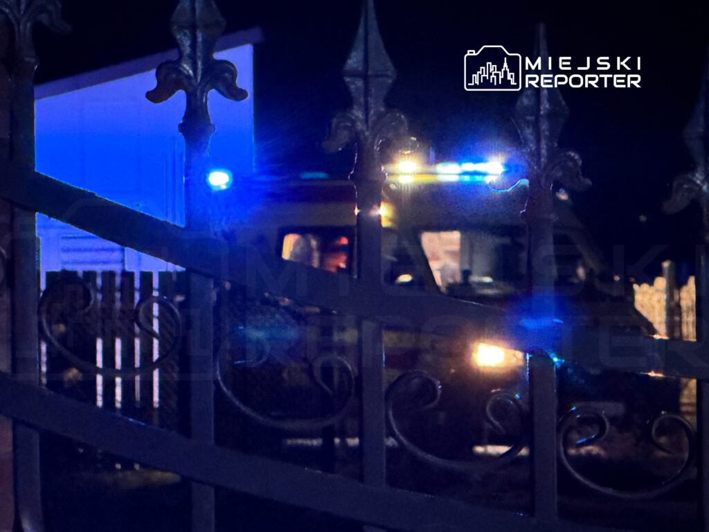 Karetka pogotowia z włączonymi sygnałami świetlnymi stoi przed ogrodzeniem w nocy.