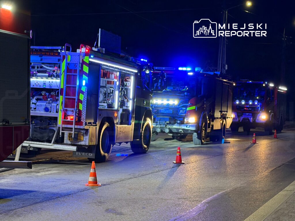 W nocy na drodze stoją dwa wozów strażackich z włączonymi sygnałami świetlnymi, otoczone pachołkami drogowymi.
