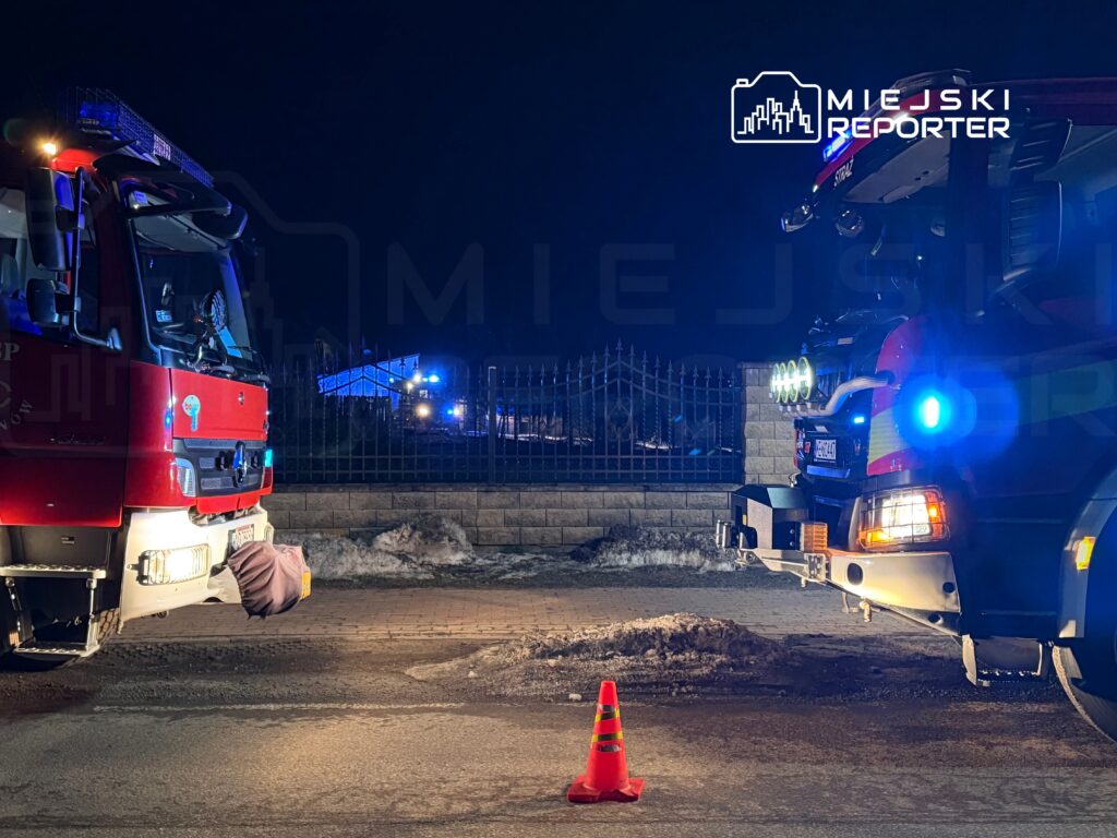 Dwa wozu strażackie zaparkowane na drodze, z niebieskimi i czerwonymi światłami w nocy, a przed nimi stoi pomarańczowy pachołek.