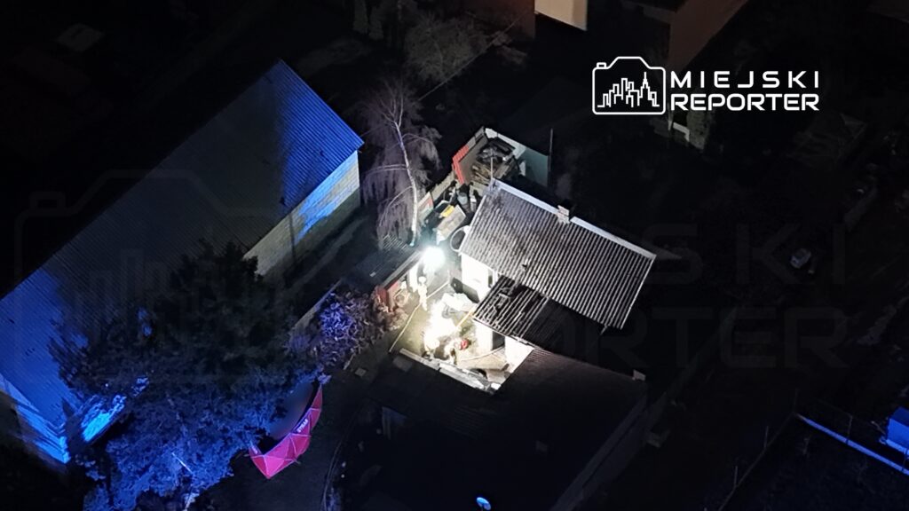 Grupa osób pracuje przy oświetlonym budynku, otoczona ciemnością i drzewami. W tle widoczne są inne budynki.