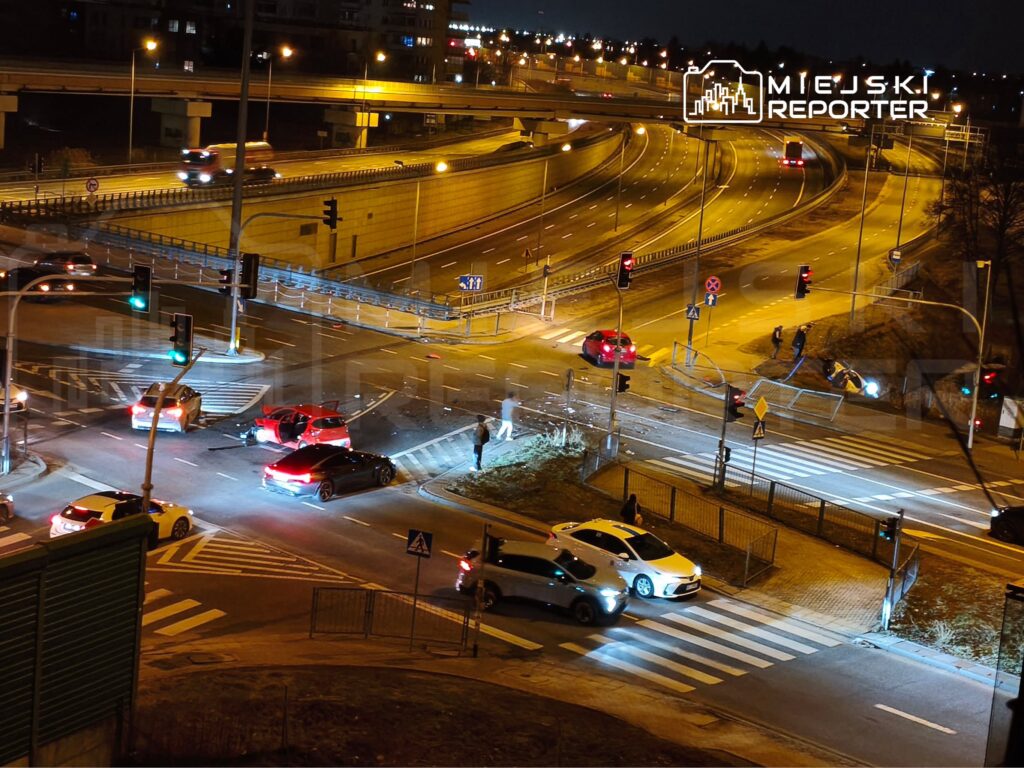 Na skrzyżowaniu ulic, kilka samochodów w tym czerwony i czarny model, uszkodzonych po kolizji, w nocy.