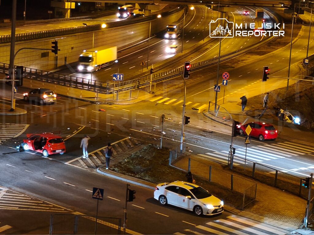 Czerwony samochód osobowy stoi na skrzyżowaniu, a obok niego kilka osób rozmawia. W tle widać inne pojazdy oraz sygnalizację świetlną.