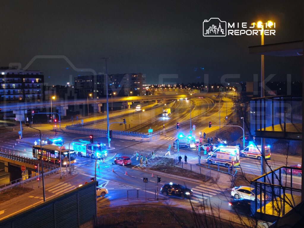 Na skrzyżowaniu z sygnalizacją świetlną stoją pojazdy ratunkowe, a obok grupka ludzi w kamizelkach odblaskowych.