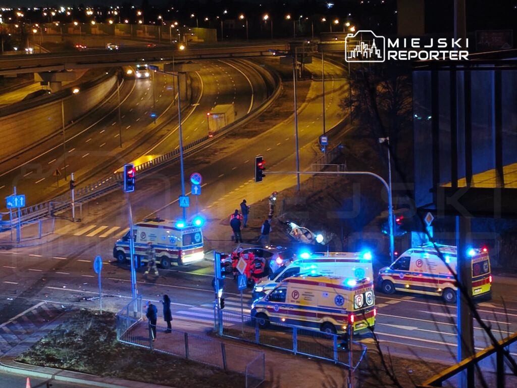 Na skrzyżowaniu ulic, przy włączonych sygnałach świetlnych, stoją karetki pogotowia, a obok grupka ludzi z interweniującymi służbami.