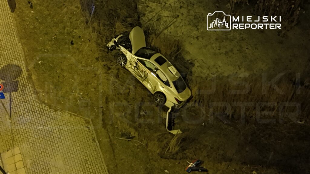 Zniszczony samochód osobowy leży w rowie, obok chodnika, otoczony trawą i śmieciami.