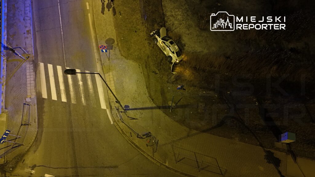 Samochód osobowy leży na boku w zaroślach przy ulicy, obok przejścia dla pieszych i oznaczenia zakazu parkowania.