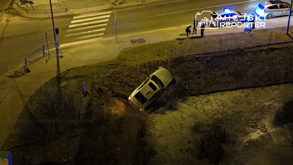 Samochód osobowy wjeżdża w row wzdłuż chodnika, obok stoją policjanci oraz kilka osób. Nocna sceneria.