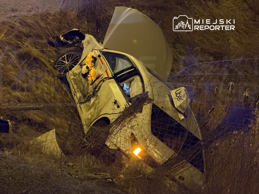 Samochód osobowy leży na boku w zaroślach przy drodze, z uszkodzoną karoserią i włączonymi światłami awaryjnymi.