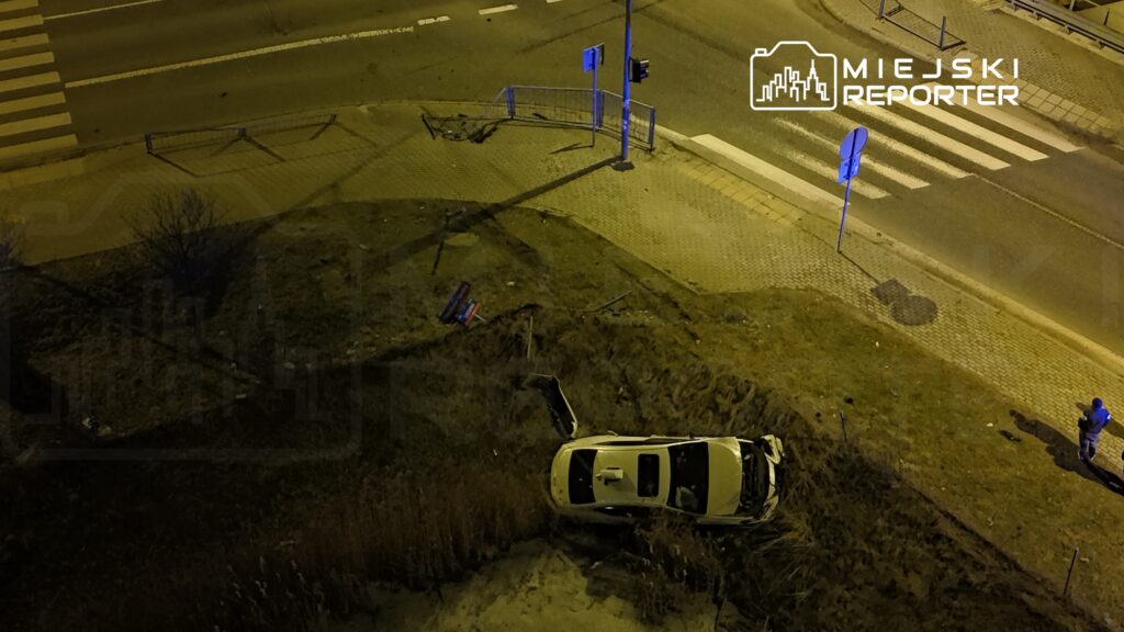 Zniszczony biały samochód leży w rowie obok jezdni, a obok stoi mężczyzna z telefonem.