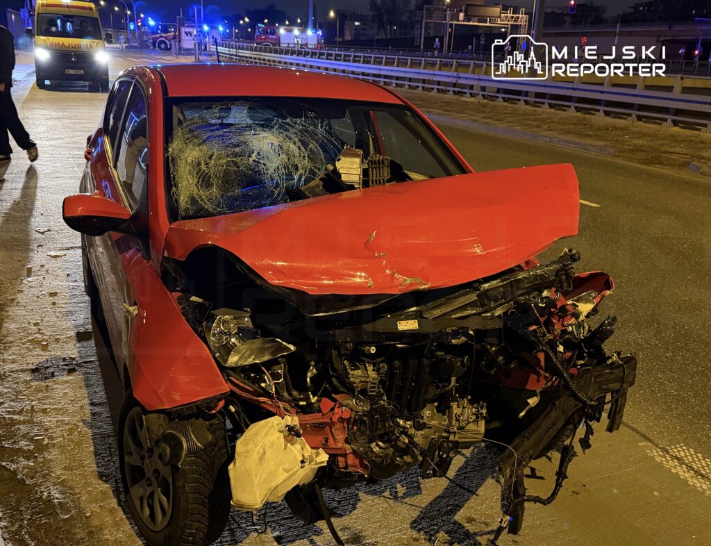 Czerwony samochód osobowy z uszkodzoną przednią częścią stoi na jezdni nocą, otoczony przez pojazdy służb ratunkowych.