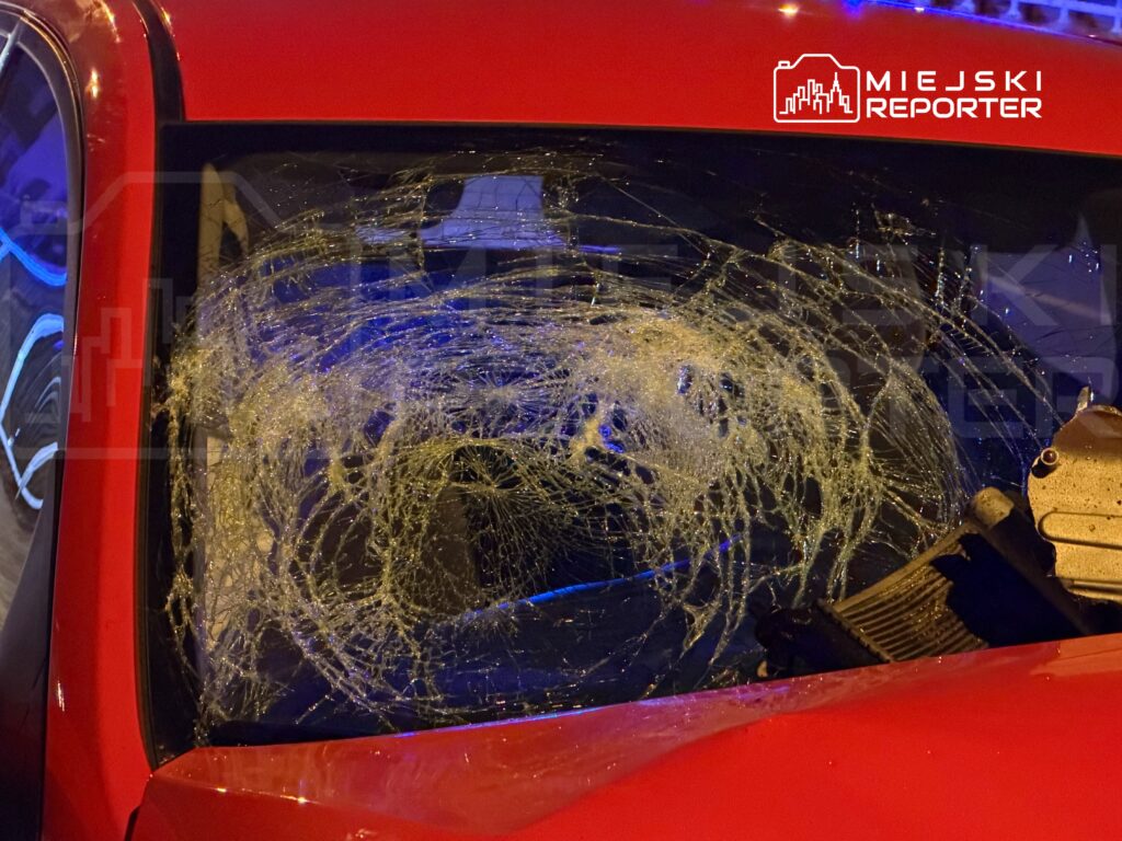 Zbity przedni szyb czerwonego samochodu z widocznymi pęknięciami, w tle niebieskie światła.