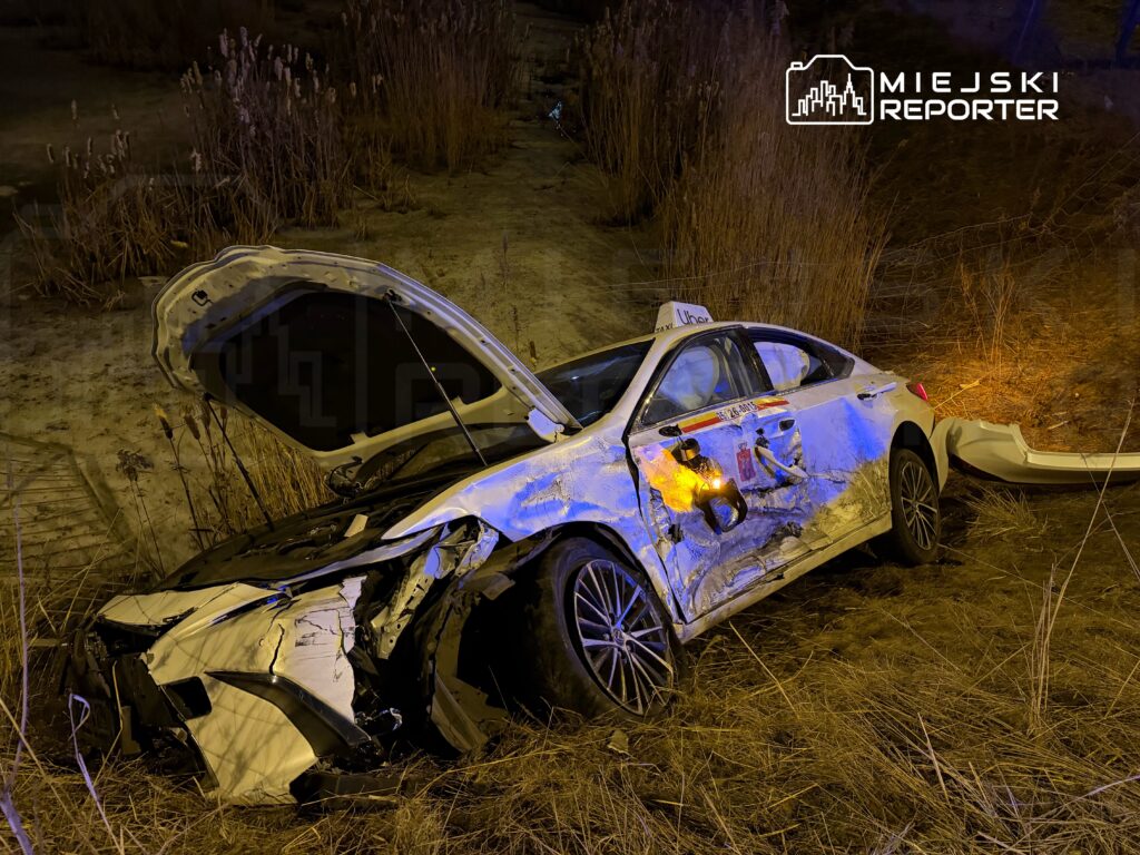 Uszkodzony samochód osobowy leży na poboczu drogi, z otwartą maską i wgniecioną karoserią, otoczony suchą roślinnością.