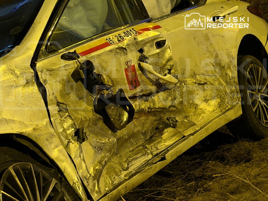 Uszkodzona strona białego samochodu osobowego z wgnieceniem na drzwiach i uszkodzoną szybą, stojącego na trawie.