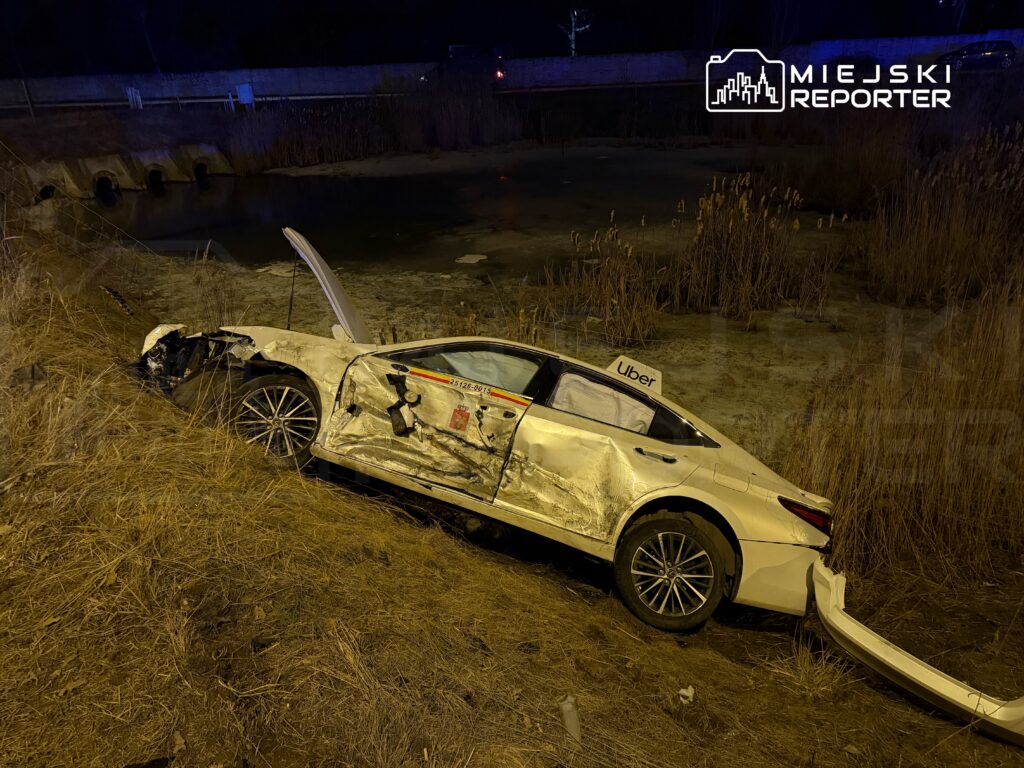 Uszkodzony samochód osobowy z logo Uber leży na zboczu w pobliżu stawu, otoczony suchą trawą.