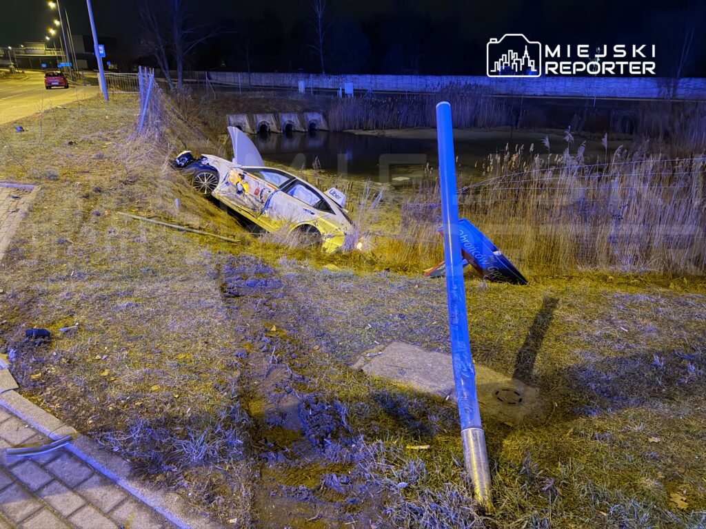 Samochód z logo Ubera wpadł do rowu przy drodze, otoczony trawą i zniszczonym oznakowaniem.