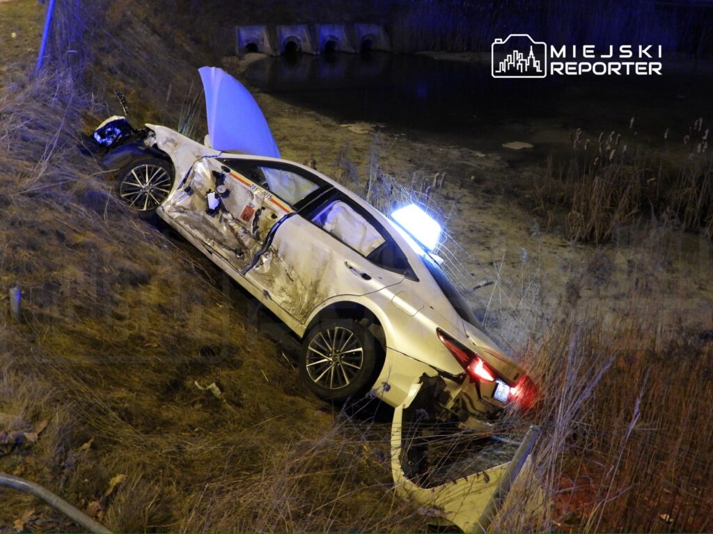 Rozbity samochód osobowy leży na poboczu drogi, częściowo w rowie na tle stawu, otoczony trawą.