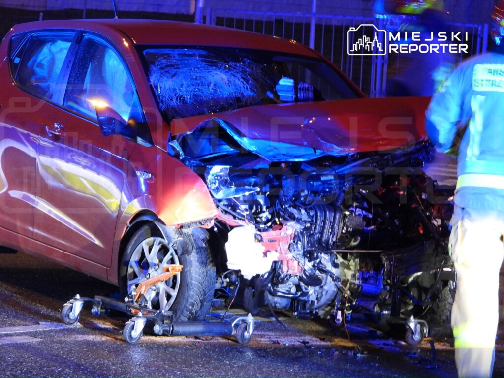 Czerwony samochód osobowy z uszkodzoną przednią częścią stoi na jezdni, obok pracują strażacy w odblaskowych strojach.