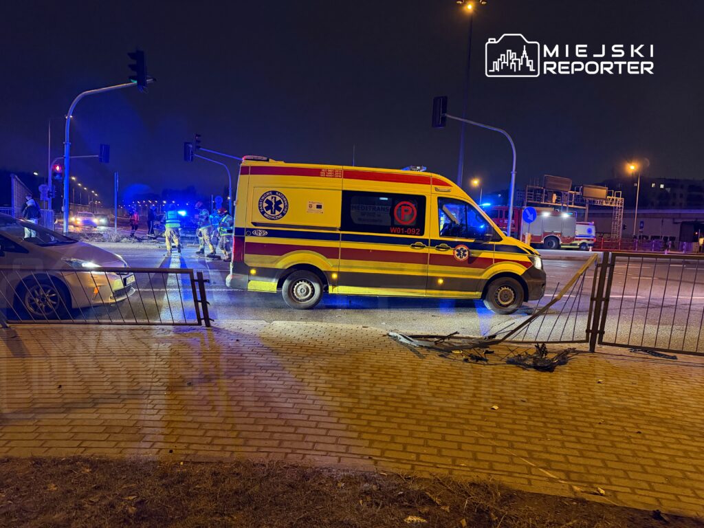 Karetka pogotowia na oświetlonej ulicy, obok grupy ratowników w strojach ochronnych, podczas nocnej interwencji.