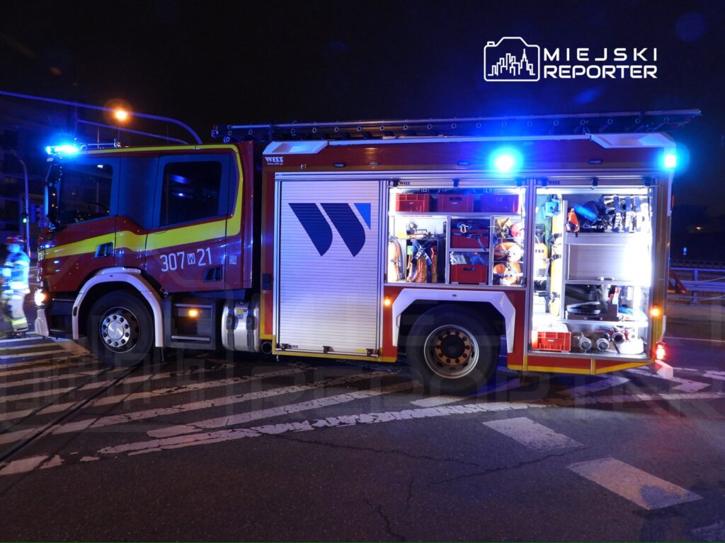 Czerwony wóz strażacki z włączonymi sygnałami świetlnymi stoi na skrzyżowaniu, otwarta szafka z wyposażeniem.