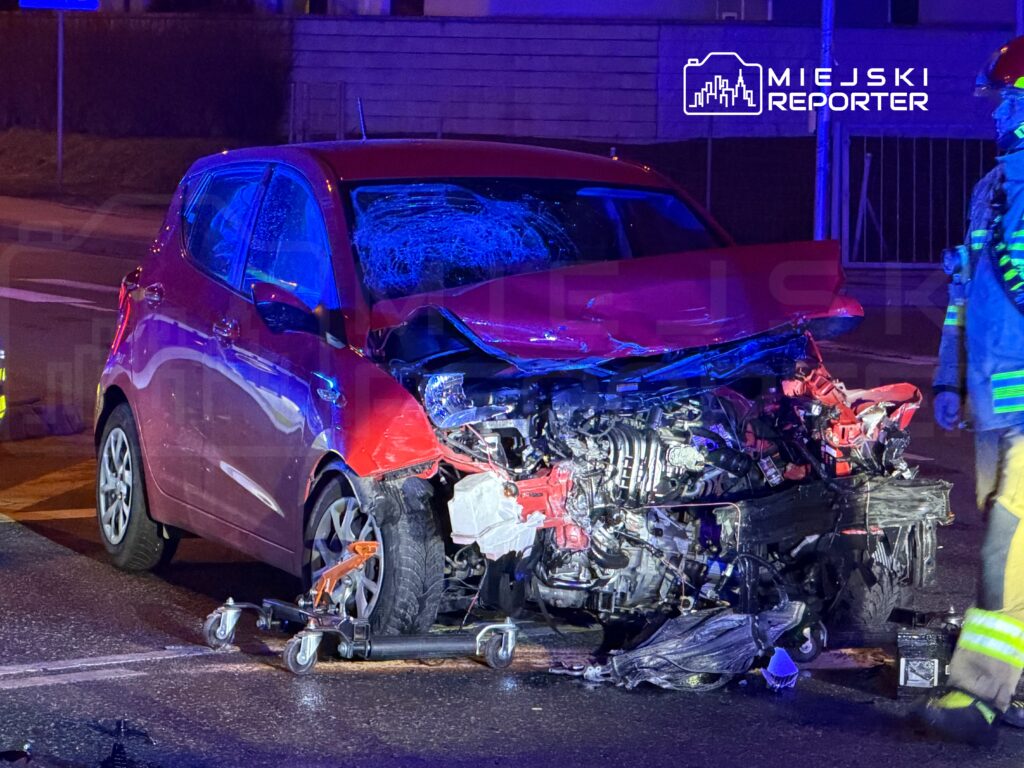 Czerwony samochód osobowy z poważnymi uszkodzeniami przodem stoi na drodze, obok strażak w mundurze.