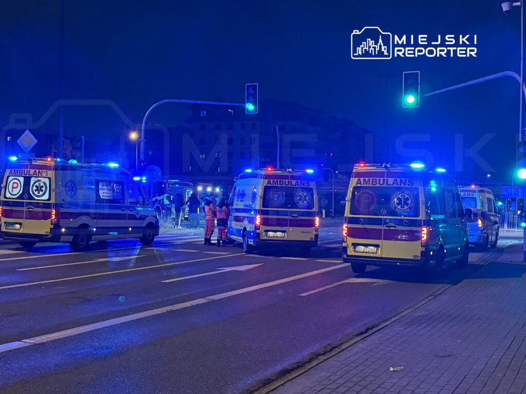 Zespół ratowników w mundurach rozmawia obok kilku ambulansów z włączonymi sygnałami świetlnymi na ulicy w nocy.