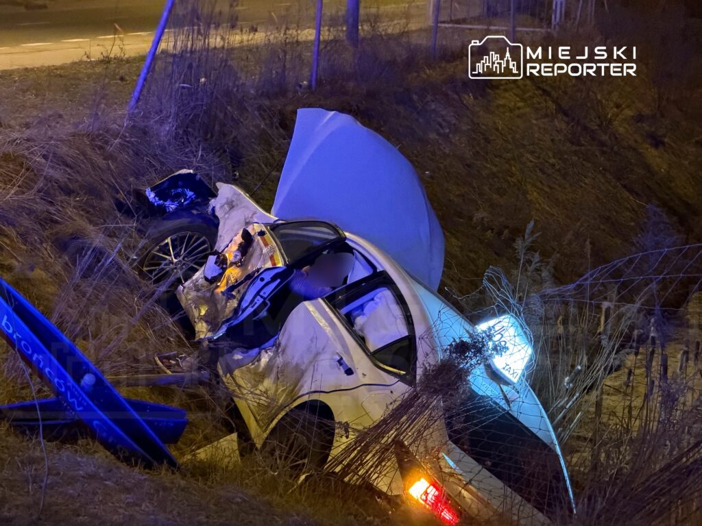 Zniszczony samochód osobowy leży na poboczu drogi, z otwartą maską i włączonym światłem.