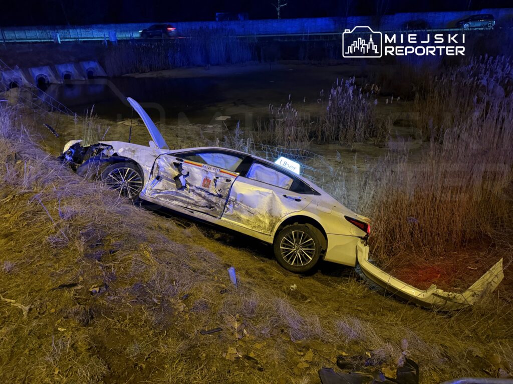 Uszkodzony samochód osobowy leży na bocznej skarpie przy drodze, z otwartą drzwiami i uszkodzoną karoserią.