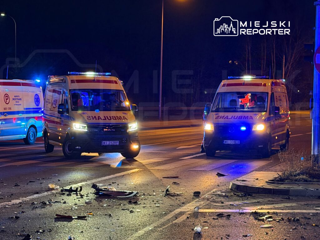 Dwa karetki pogotowia z włączonymi sygnałami świetlnymi zatrzymane na skrzyżowaniu, w tle widoczne fragmenty wraku pojazdu.