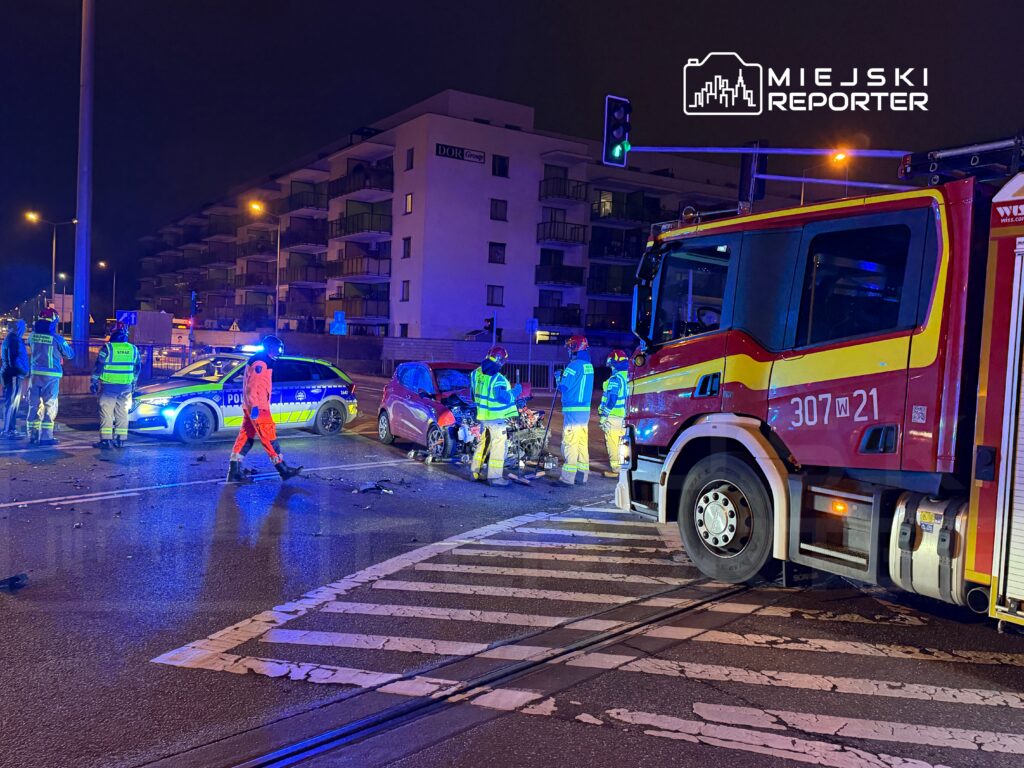 Ratownicy i policjanci pracują na miejscu wypadku drogowego, obok uszkodzonego samochodu oraz wozu strażackiego.