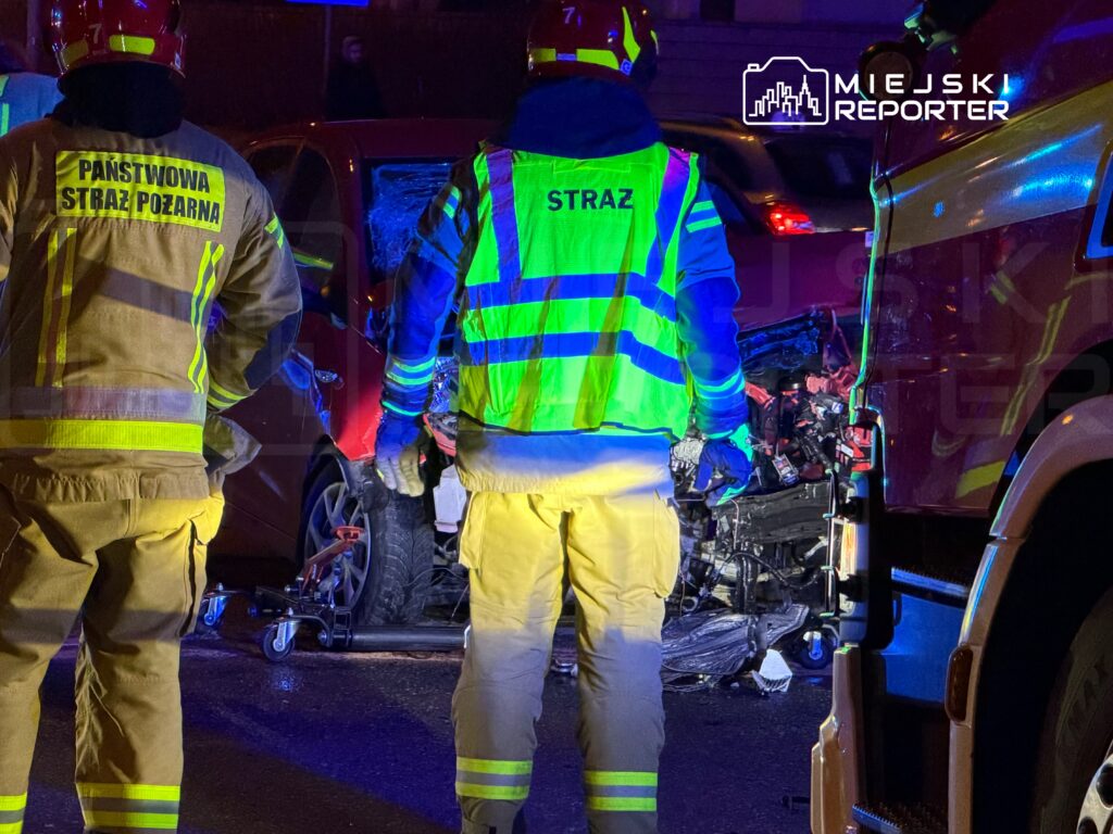 Strażacy w odblaskowych strojach pracują przy wypadku drogowym, obok uszkodzonego samochodu osobowego.