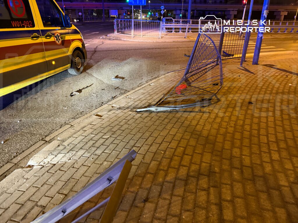 Uszkodzona barierka leży na chodniku obok parkującego pojazdu służb ratunkowych w nocnym otoczeniu miejskim.