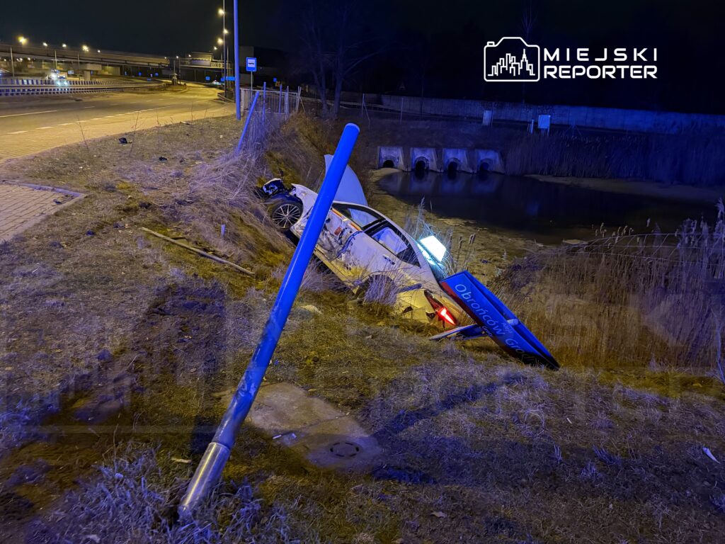Samochód wypadł z drogi i utknął w rowie obok stawu, obok leży niebieski znak drogowy. Nocna sceneria.