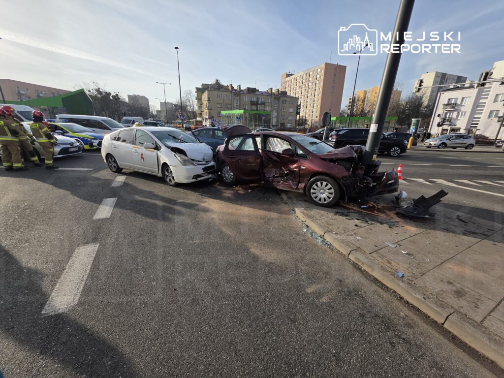 Dwa uszkodzone samochody osobowe zderzone na skrzyżowaniu, obok stoją strażacy i policja.