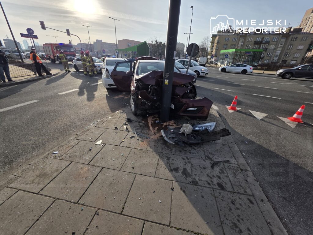 Czerwony samochód osobowy uderzył w latarnię, na chodniku leżą odłamki szkła, a obok pracują strażacy.