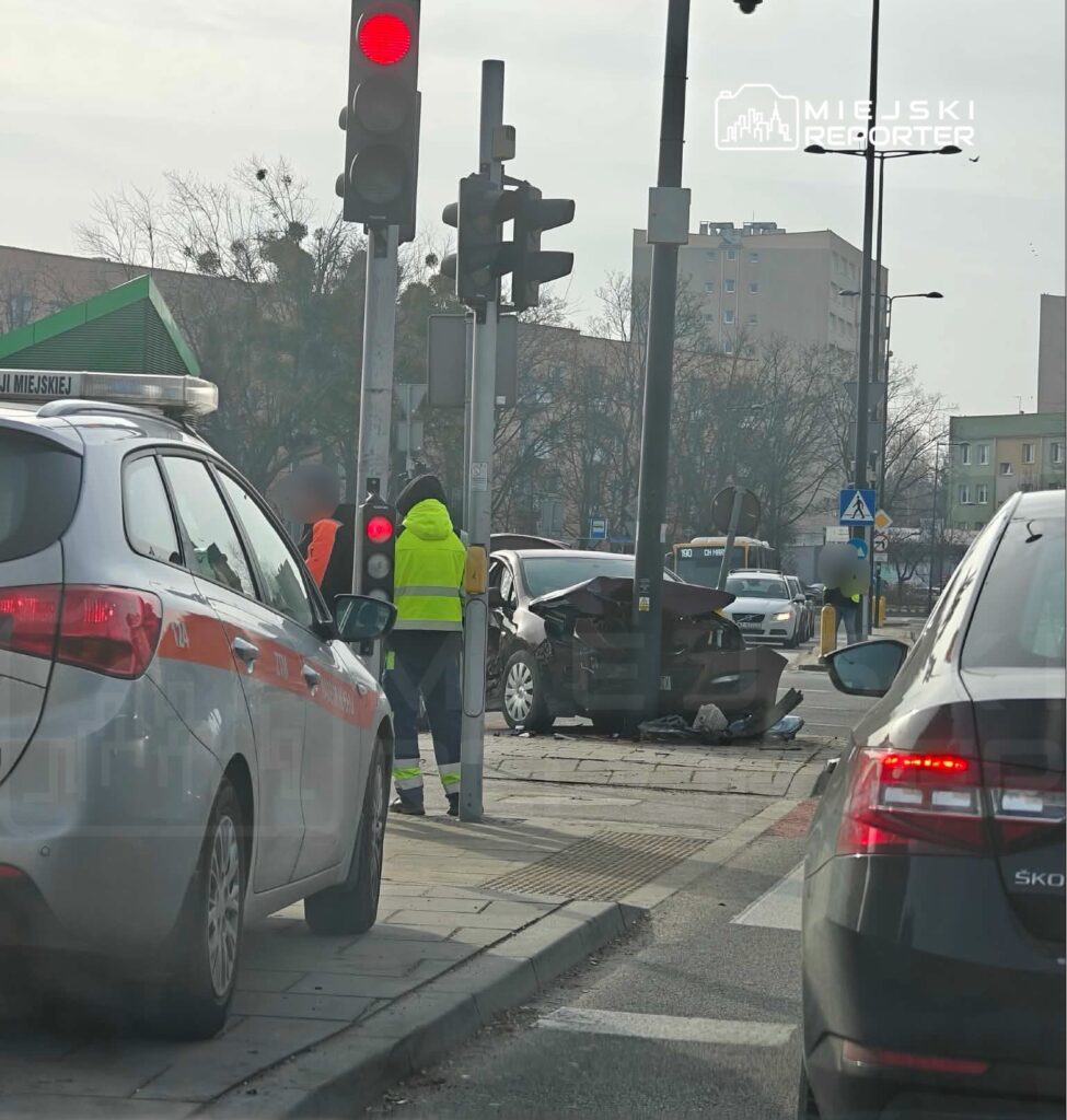 Na skrzyżowaniu dwóch ulic, ratownicy w odblaskowych kamizelkach oceniają skutki wypadku samochodowego.