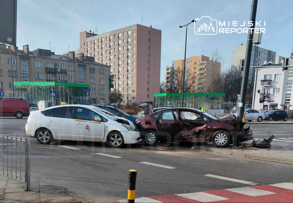 Dwa uszkodzone samochody stoją na skrzyżowaniu, w tle widoczne budynki mieszkalne i zielona przestrzeń.