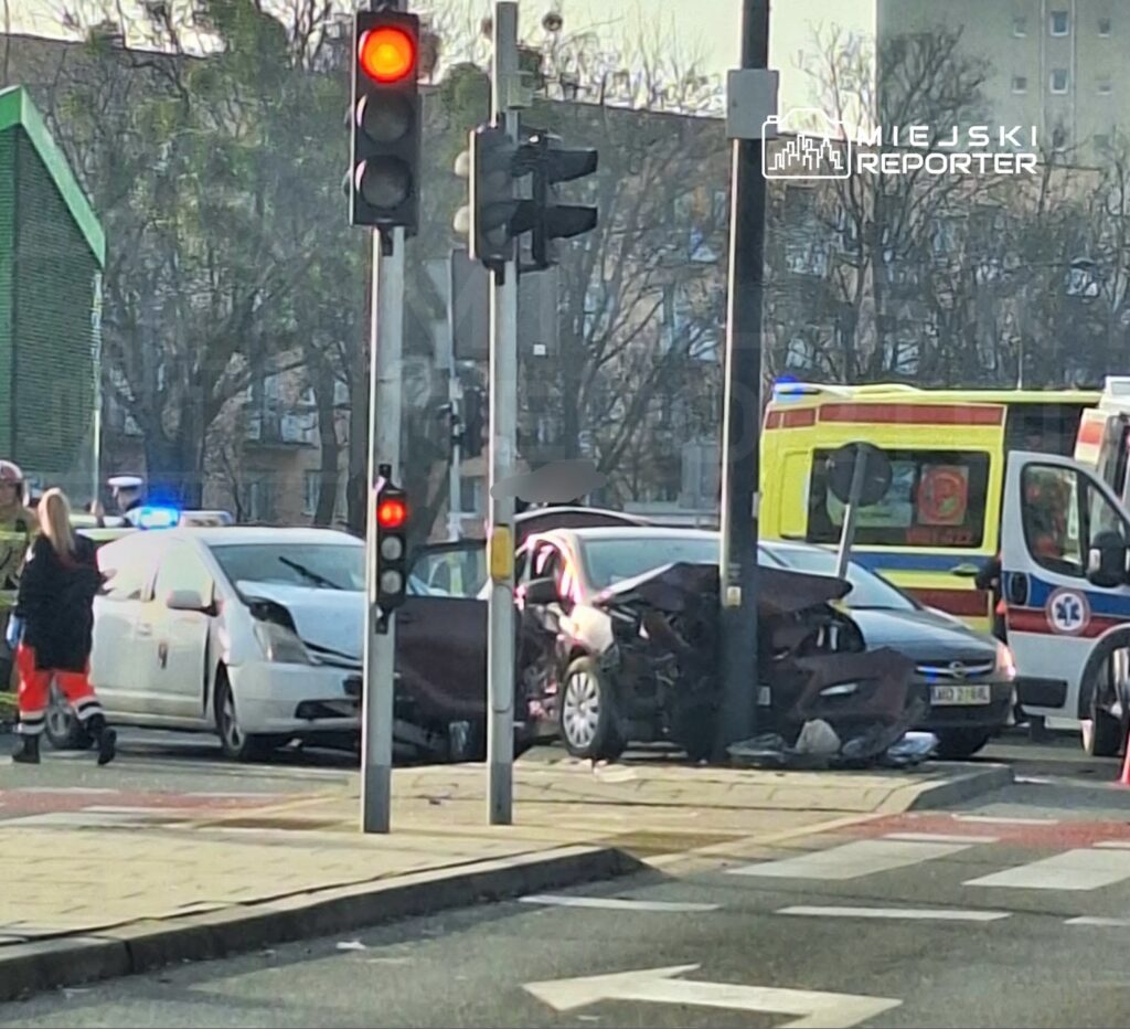 W wyniku wypadku drogowego dwa uszkodzone samochody stoją na skrzyżowaniu, a obok interweniują służby ratunkowe.
