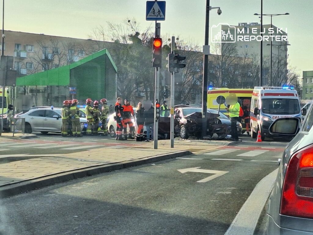 Grupa strażaków i ratowników medycznych udziela pomocy po wypadku drogowym na skrzyżowaniu, obok uszkodzonych pojazdów.