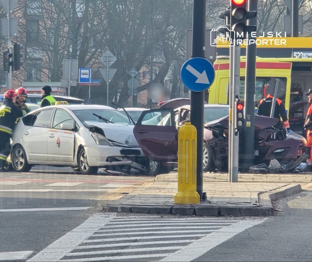 Na skrzyżowaniu stoją uszkodzone samochody, wokół których pracują strażacy i ratownicy medyczni.