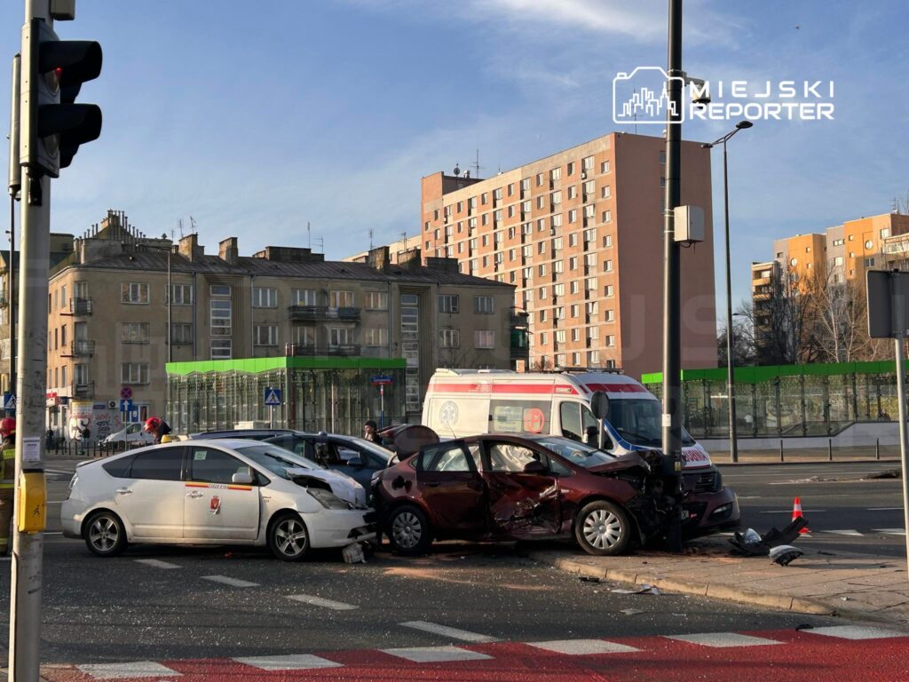 Dwa uszkodzone samochody po zderzeniu na skrzyżowaniu, obok stoi karetka pogotowia, w tle budynki mieszkalne.