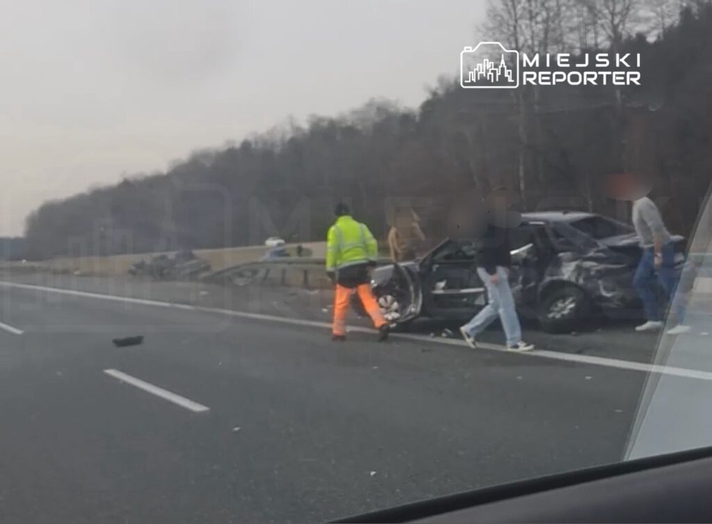 Na autostradzie grupa osób zbiera się wokół uszkodzonego samochodu, a pracownik w odblaskowej kamizelce kieruje ruchem.