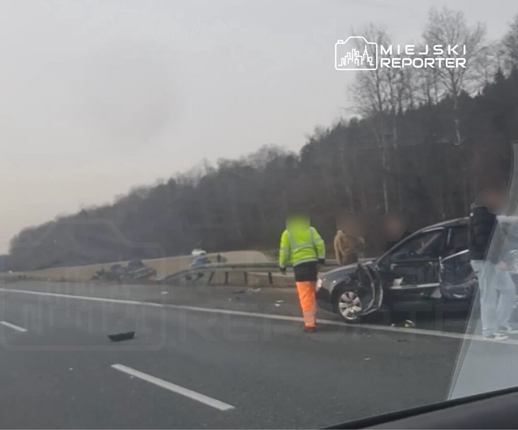 Na drodze leży uszkodzony samochód, a obok niego stoi mężczyzna w żółtej kamizelce i kilku innych ludzi.