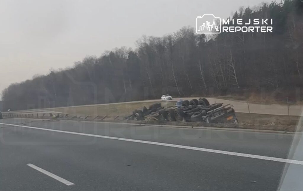Zniszczony samochód ciężarowy leży na boku obok drogi, a w tle widać pojedynczy samochód osobowy.