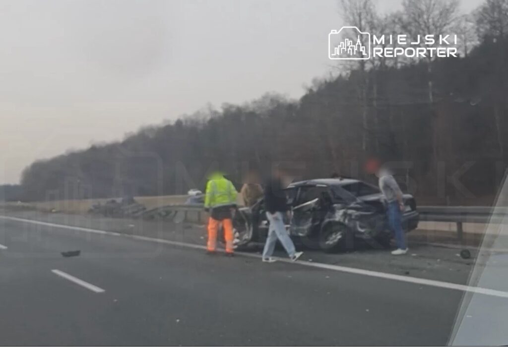 Grupa osób w odblaskowych kamizelkach ocenia uszkodzenia samochodu po wypadku na autostradzie.
