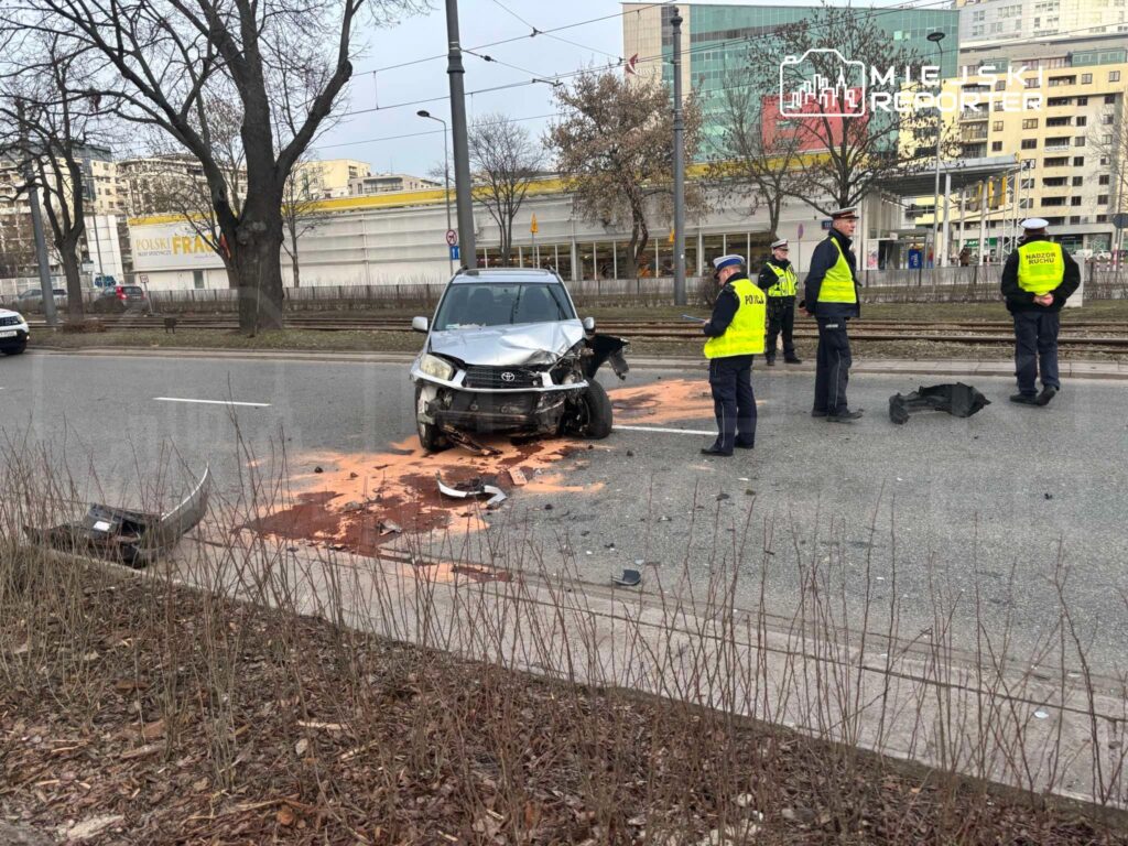 W miejscu wypadku drogowego, policjanci w żółtych kamizelkach zabezpieczają teren wokół uszkodzonego samochodu.