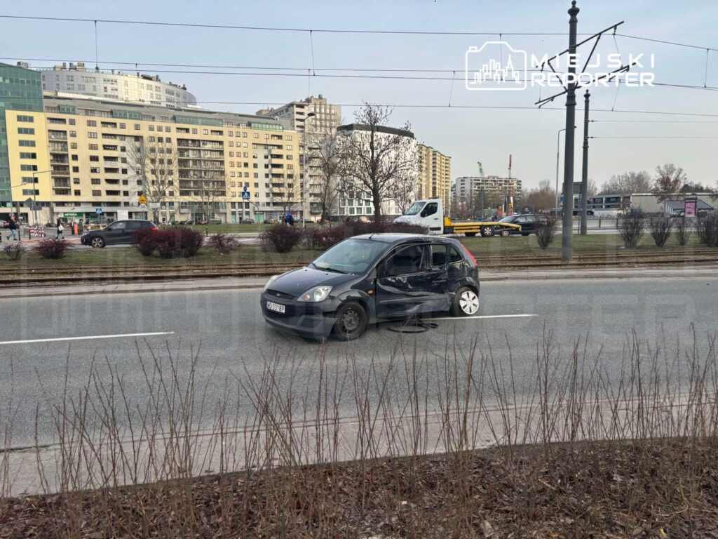 Uszkodzony czarny samochód osobowy stoi na jezdni obok nowoczesnych budynków mieszkalnych.