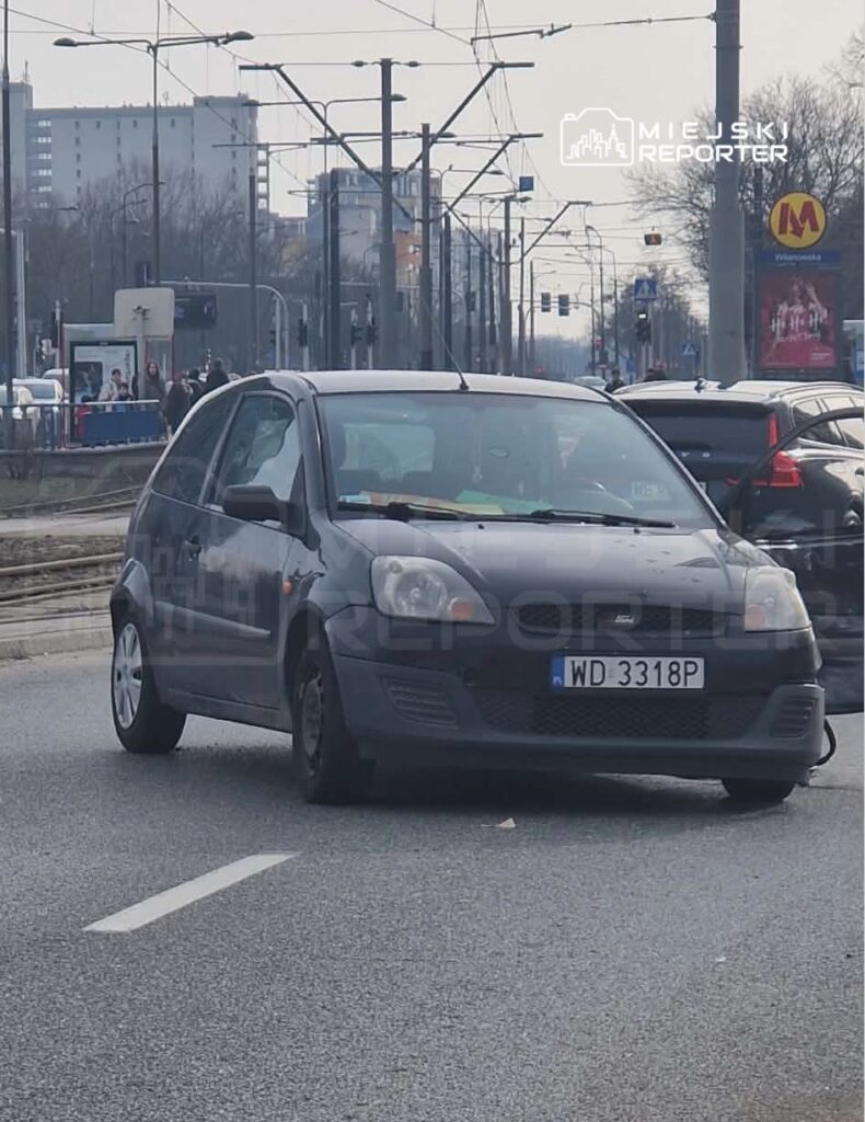 Czarny samochód osobowy porusza się po miejskiej ulicy w otoczeniu innych pojazdów.
