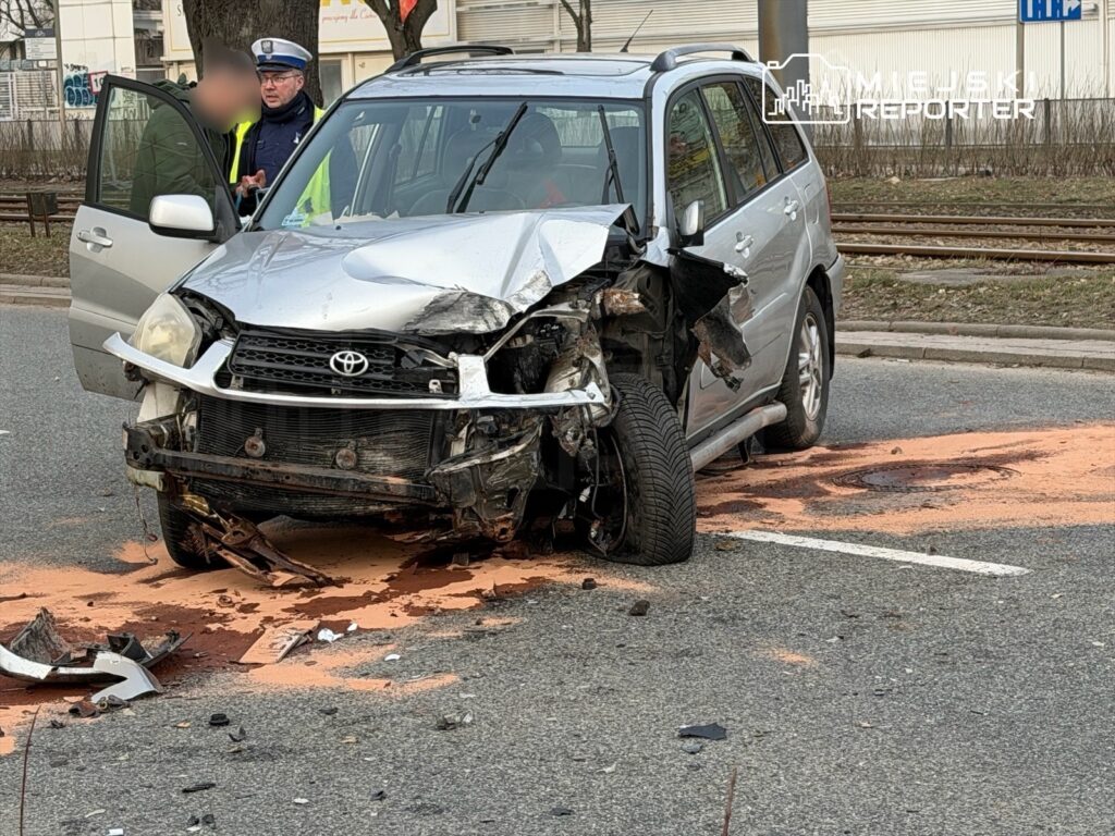 Rozbity samochód osobowy na jezdni, obok policjant rozmawia z kierowcą. Na drodze widoczne plamy płynów eksploatacyjnych.