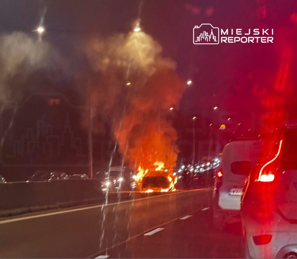Pojazd w płomieniach na pasie ruchu, obok stojące samochody w korku, nocna sceneria oświetlona latarniami.